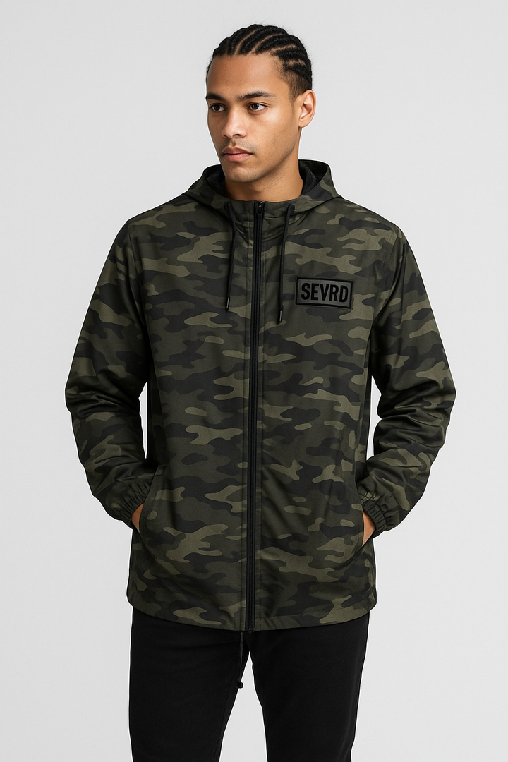 Model wearing SEVRD forest camo windbreaker jacket with black embroidered box logo patch, standing in an urban setting. Features snap front, drawcord hem, and tactical fit