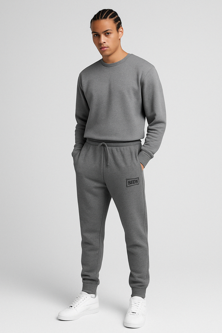 Model wearing SEVRD Stamp Joggers in rebel gray fleece with black embroidered box logo, standing in minimalist urban setting. Streetwear fit, ribbed cuffs, and deep pockets visible.